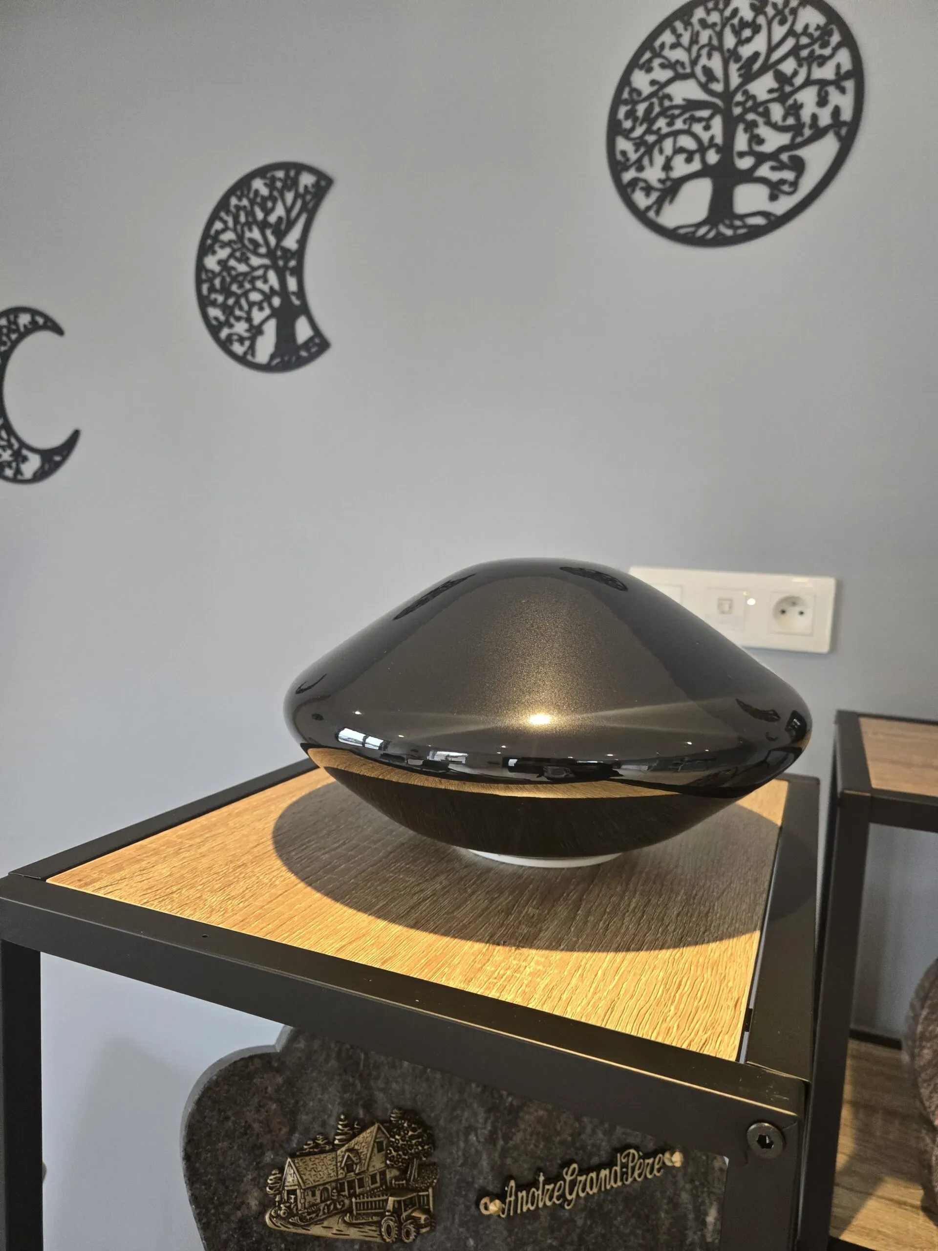 Urne cinéraire moderne en forme de galet brillant gris foncé/anthracite posée sur un meuble en bois.